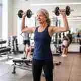 A fit 72-year-old woman doing a standing dumbbell shoulder press in a bright modern gym