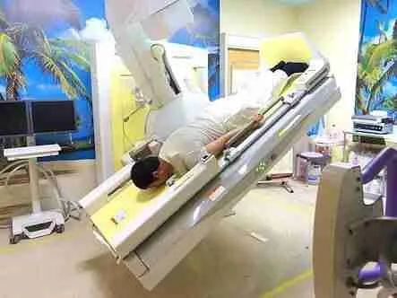 A man lying face down on a Japanese barium X-ray table tilted steeply head-down, in a room decorated with palm tree wallpaper